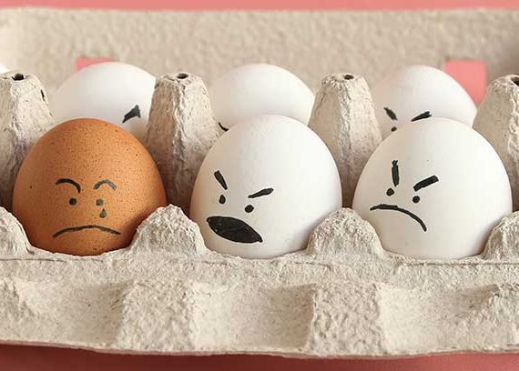 Group of white organic chicken eggs with angry faces and one brown chicken egg with crying face in carton box made from recycled paper.