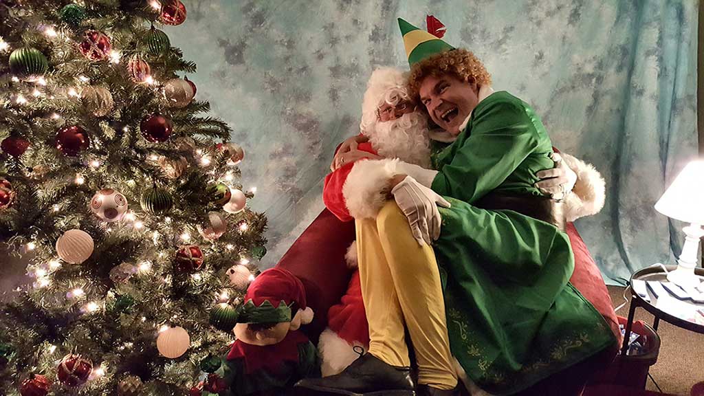 Elf sitting on Santa's Lap next to a Christmas Tree
